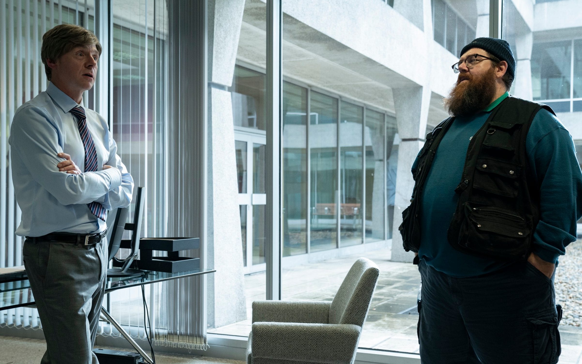 Gus speaks to his boss, Dave, in his office. They are both standing. Dave is in a collared shirt and a tie while Gus is in his beanie and sweatshirt with a black vest.