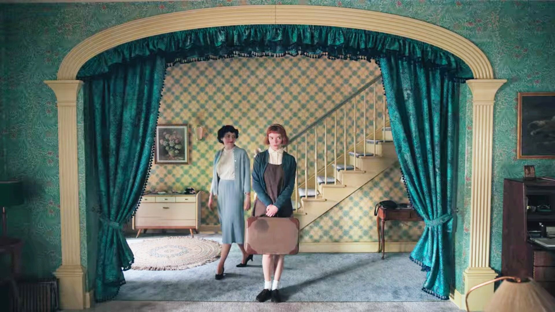 Mrs. Alma Wheatley (Marielle Heller) and Beth Harmon (Anya Taylor-Joy) stand in front of the staircase of the Wheatley home with turquoise floral wallpaper and turquoise satin curtains.
