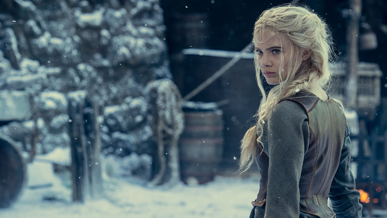 A woman looks over over her shoulder at something off-screen. In the background is a stone structure and snow.