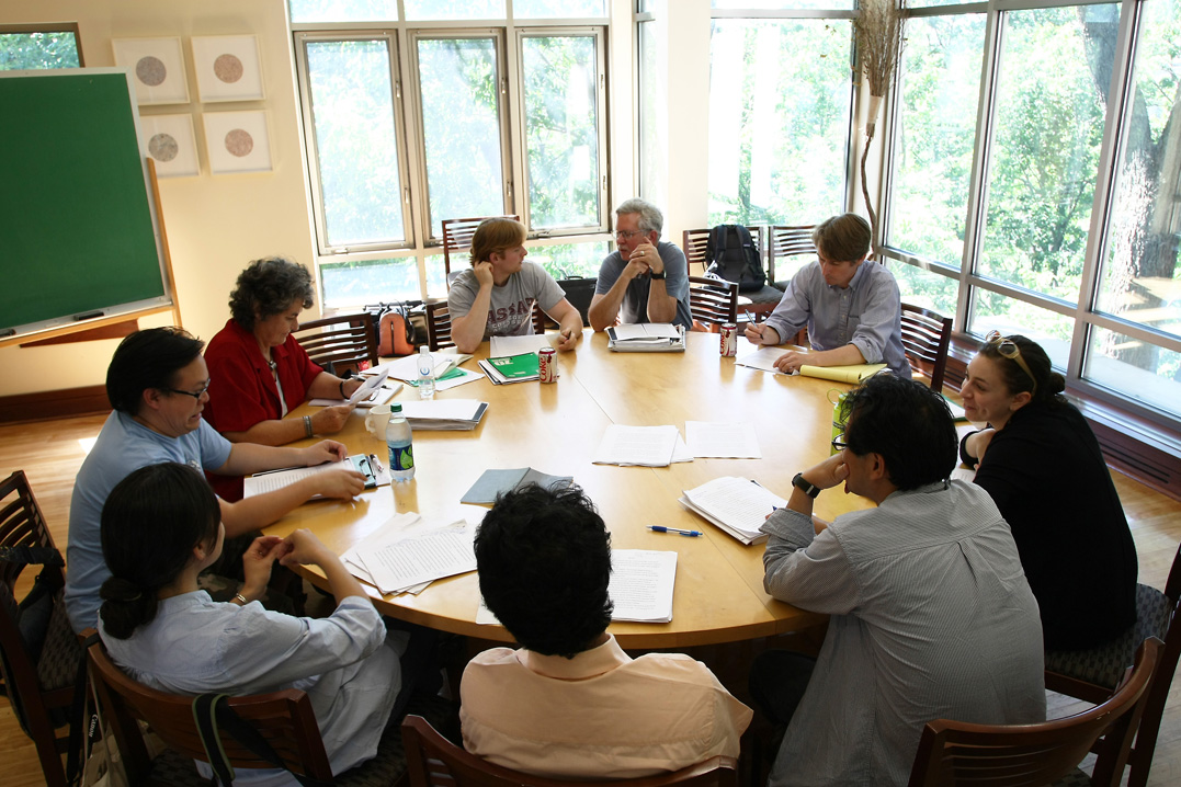 A group of writers sit around a round table talking with each other over open notebooks.