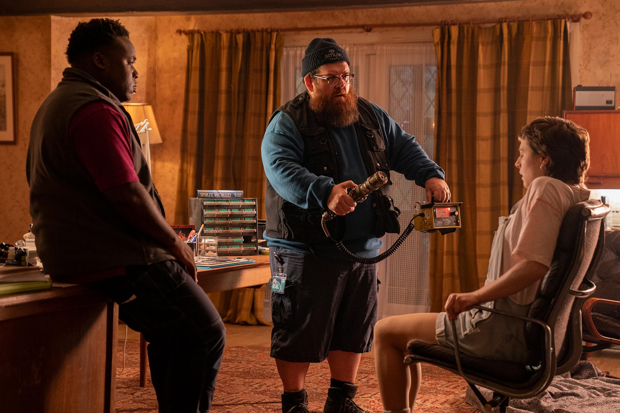 Gus using a home made "ghost detector" which looks like a microphone hooked up to an electronic box. He is scanning Astrid who is sitting in a chair. Elton watches from a few feet away. 