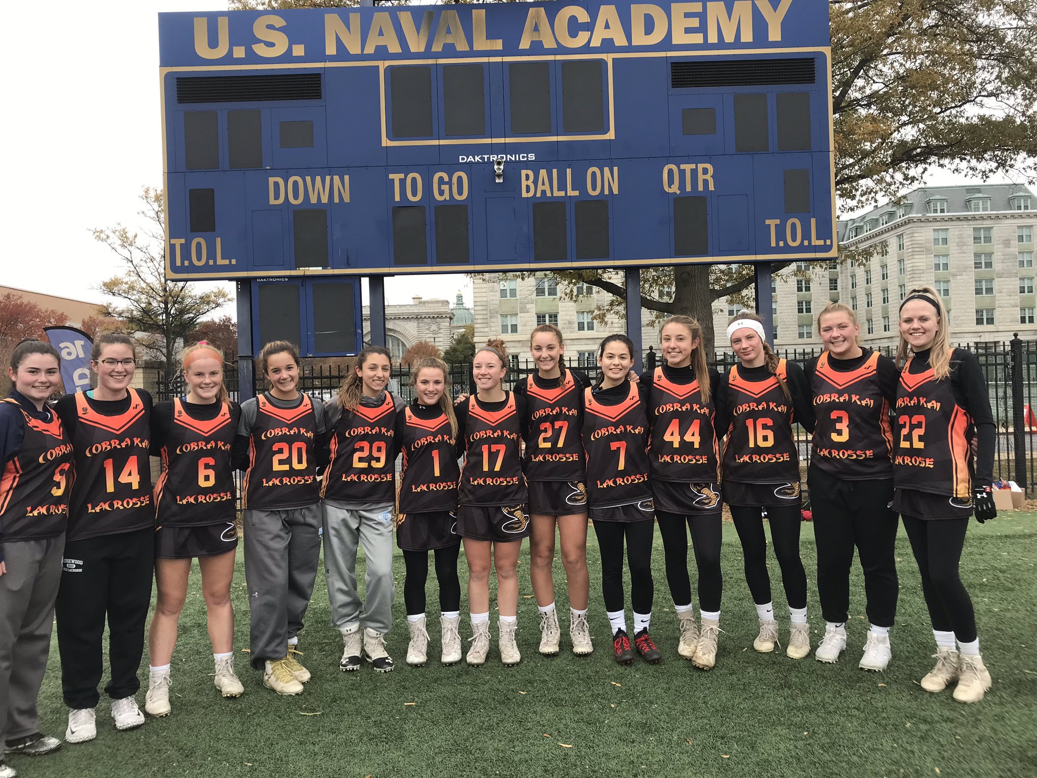 Cobra Kai 2021 team dressed in black uniforms stand in front of a Naval Academy scoreboard. 