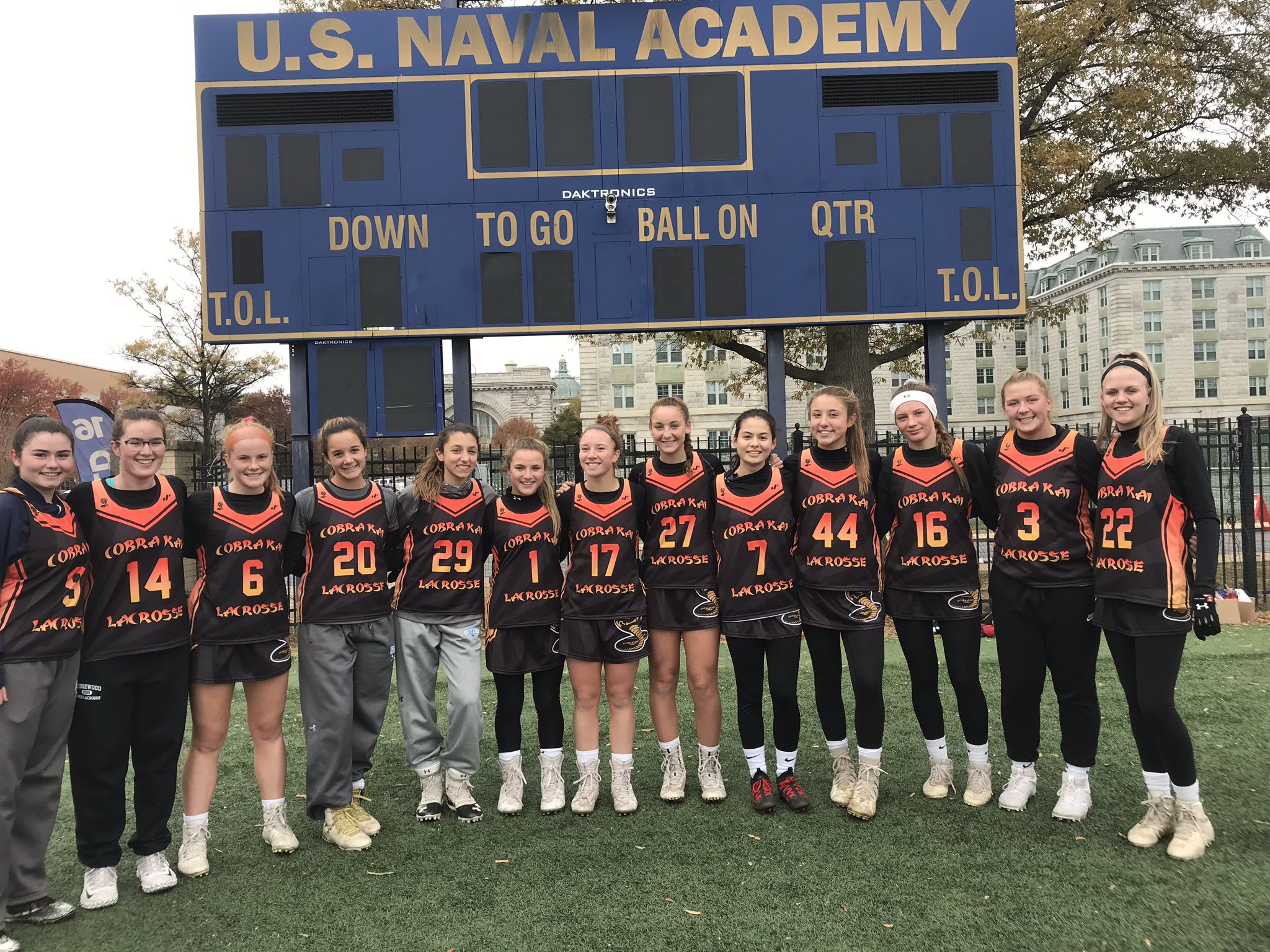 Cobra Kai 2021 team dressed in black uniforms stand in front of a Naval Academy scoreboard.