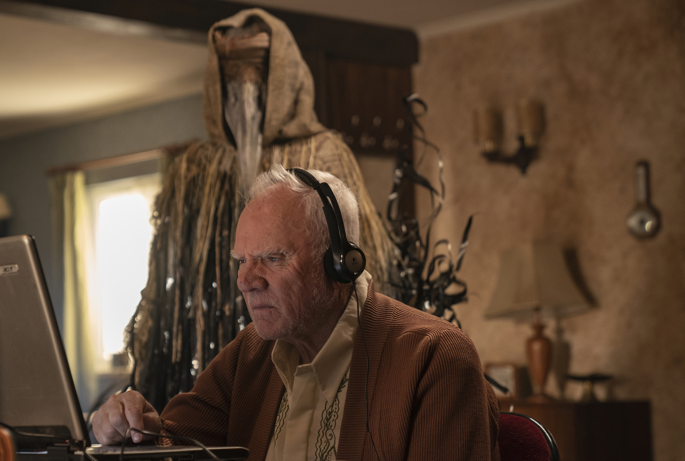 Richard, played by Malcom McDowell, sits and stares at a laptop with headphones on. He is oblivious to the person or thing standing behind him. It is shaped like a person but draped completely in garment. 