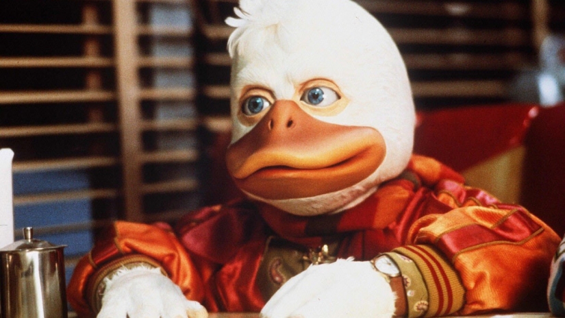 Still image of Howard the Duck in a red jacket sitting in a diner from the live-action 1986 film. The actor playing Howard the Duck is in a full-body costume. 