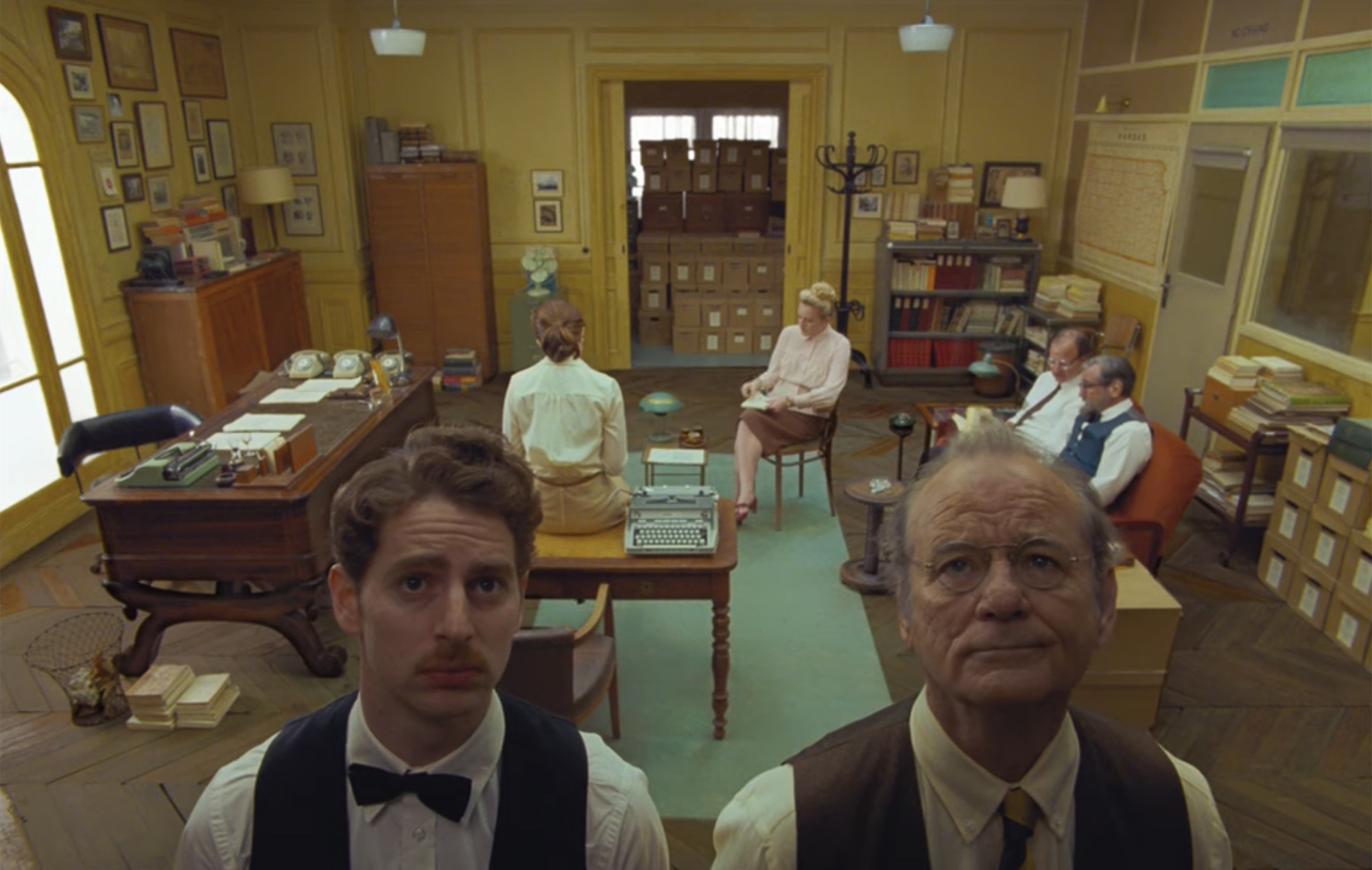 Two men look up at something just above the viewer. In the background, four other people sit in chairs in a secretarial office.