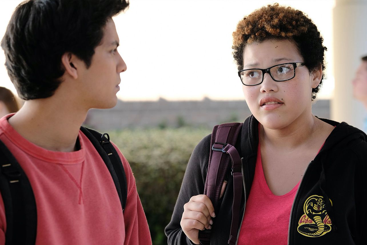 Aisha and Miguel from TV show Cobra Kai talk to each other. Both are wearing red shirts, and Aisha has on a Cobra sweatshirt. 