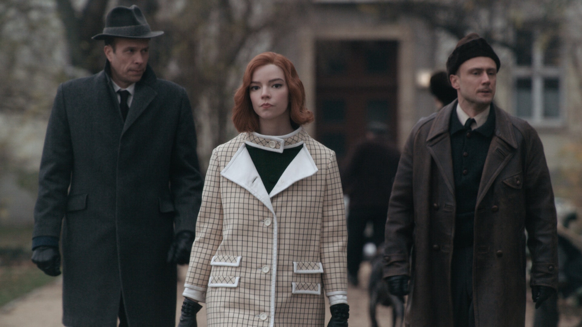 Beth Harmon (Anya Taylor-Joy) accompanied by two older men walks the streets of a city dressed in winter coats.