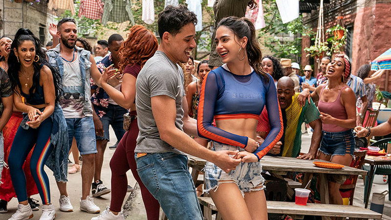 A couple smiles and dances together. In the background, a group of people dance and clap.