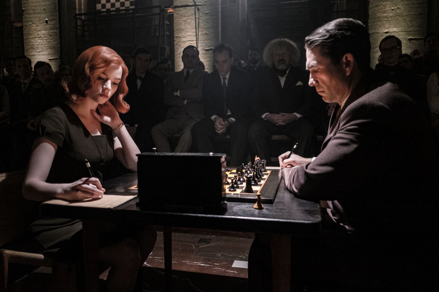 Beth Harmon (Anya Taylor-Joy) and Vasily Borgov (Marcin Dorociński) play their final game of chess in a dark Russian basement, while Beth appears to have an expression that says "unbothered" and Borgov looks nervous.