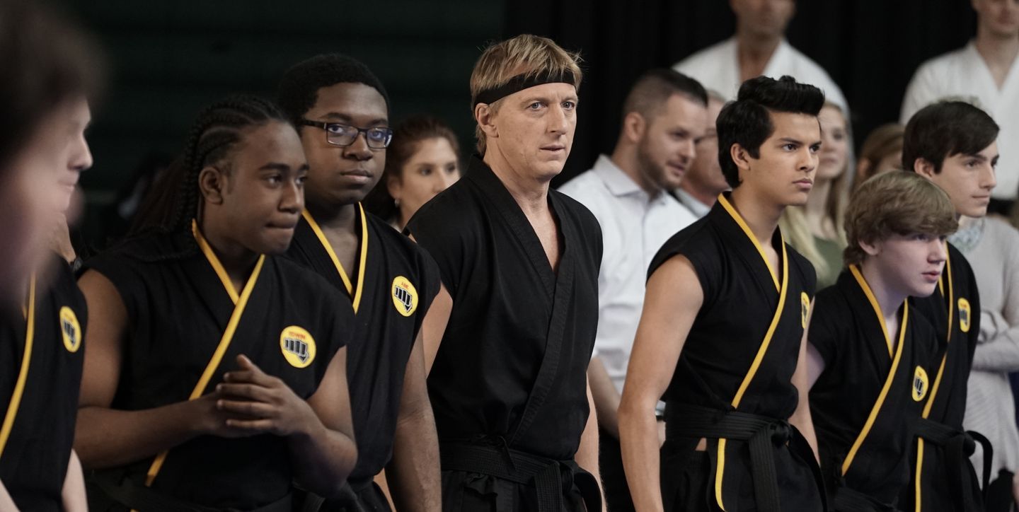 Johnny stands in the center of his Cobra Kai students. Everyone is dressed in black uniforms. 