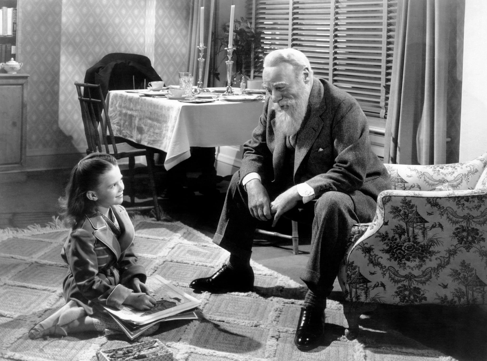 Susan Walker, seated on the floor, looks up at Kris Kringle, seated in a chair. Both are smiling at each other.