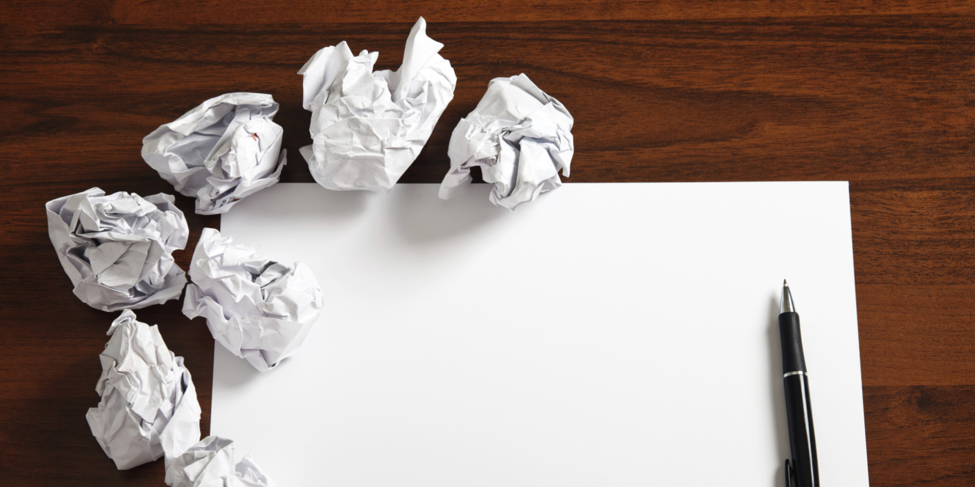 A blank paper and pen sit on a table with several wadded up pages to the side.