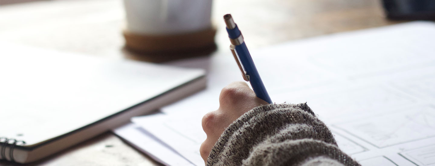 A person writes with a pen on papers strewn on a table.