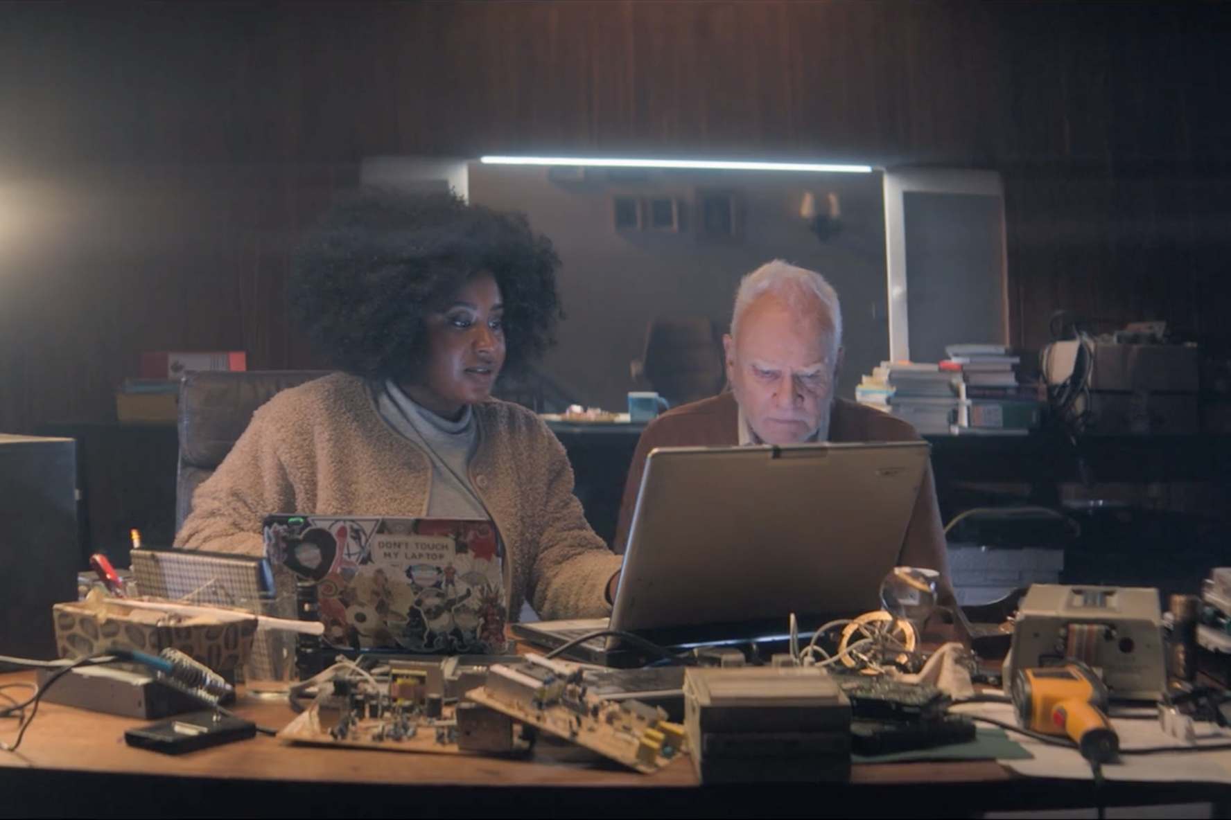 Helen sits with Richard at a desk strewn with electronic equipment. They both have laptop computers open as they stare at Richard's screen.