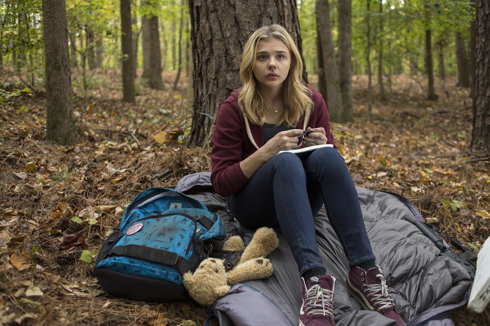 Cassie Sullivan, protagonist of The Fifth Wave, is potrayed by Chloe Grace Moretz. She sits, anxious, curled up on a sleeping bag in the woods with a teddy bear at her feet.