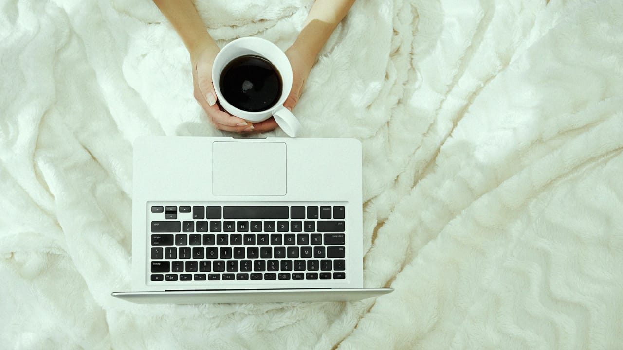 A person sits in bed with a cup of coffee and a laptop.