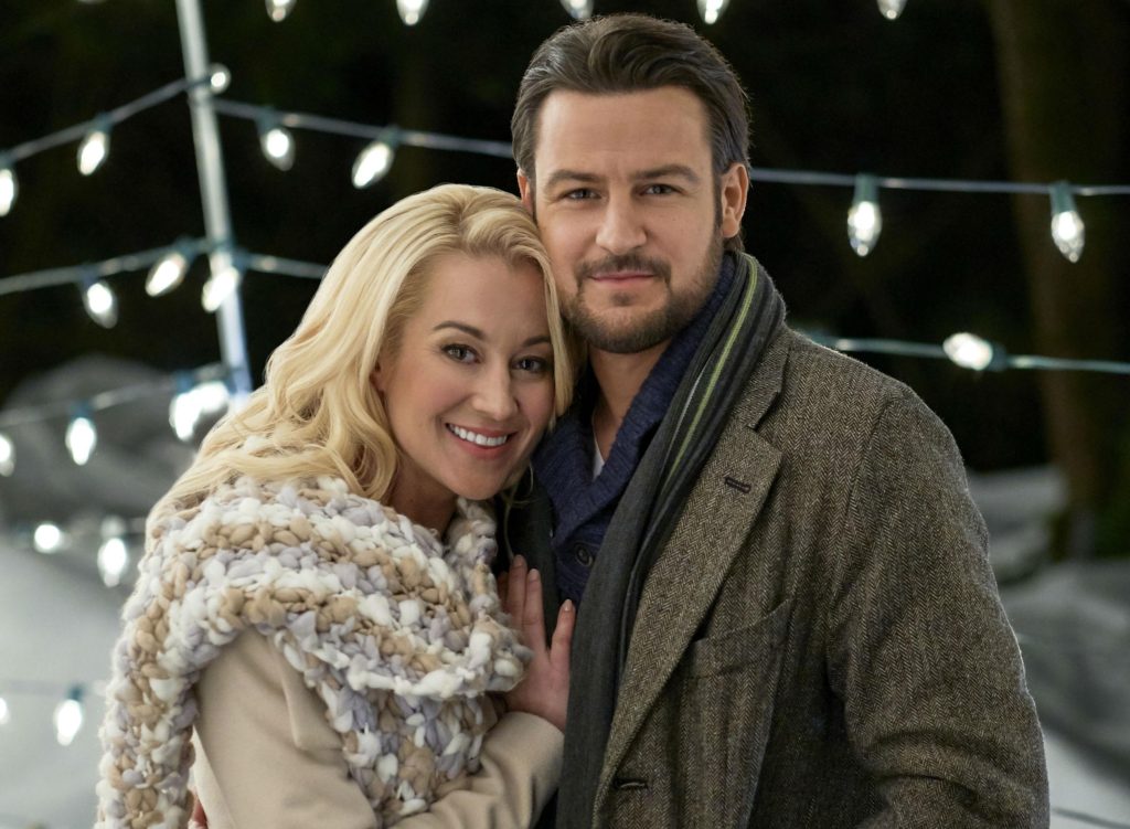 The main couple stands close together while in front of Christmas lights.