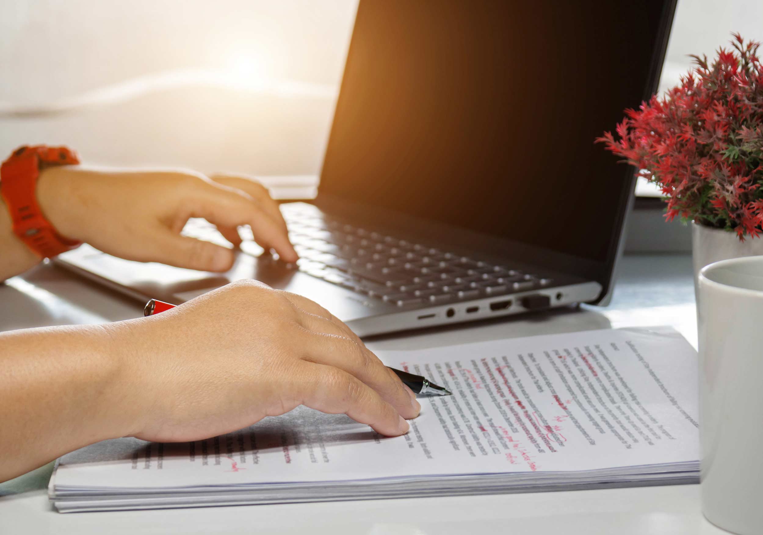 A person edits a printed out manuscript with their laptop beside the paper.