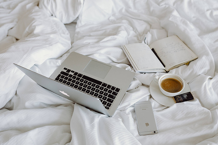 A laptop, notebook, cell phone, and coffee cup sit on a bed.