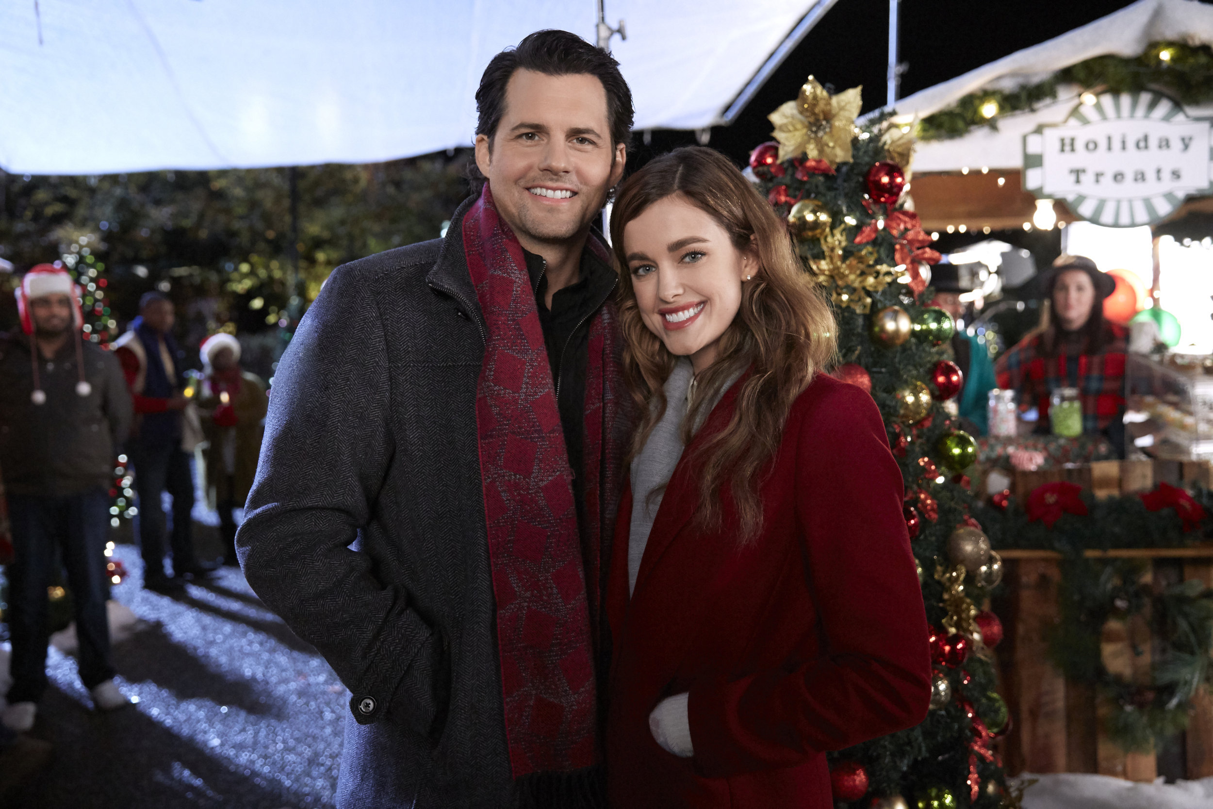 The two smiling main characters stand close together while at an outdoor Christmas market.