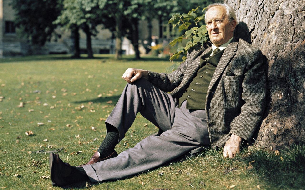  J.R.R. Tolkien wears a grey tweed suit and dark green vest while sitting leisurely in the grass leaning against a tree trunk.