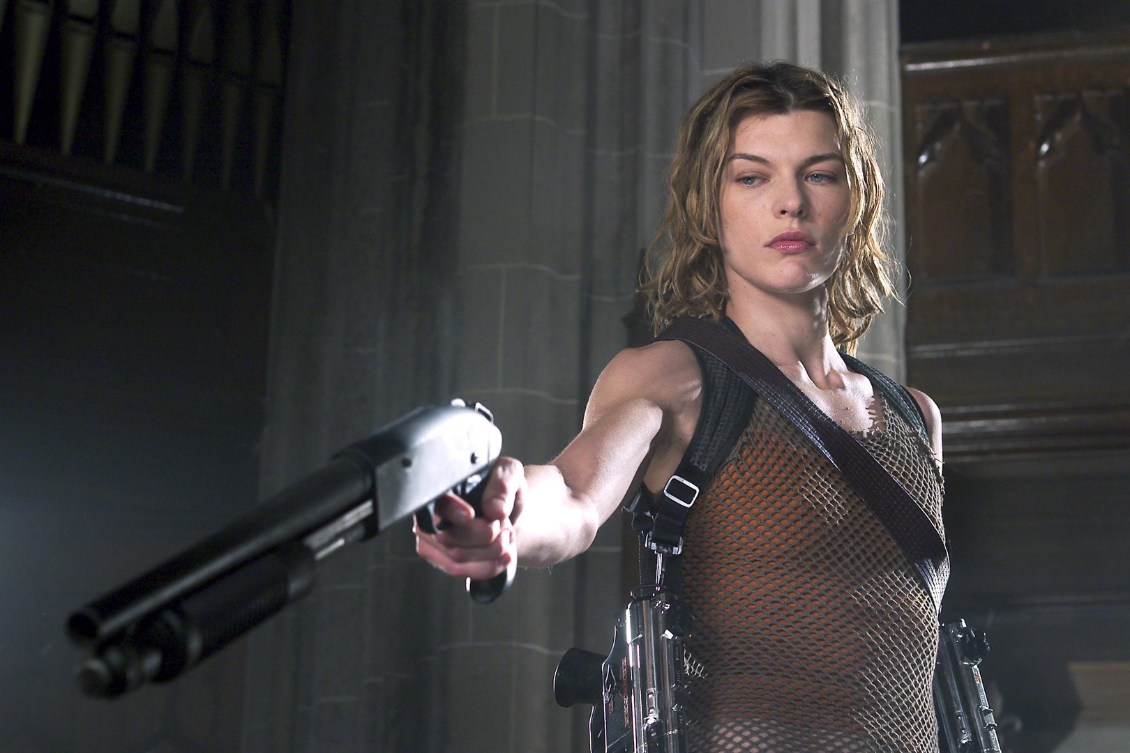 Milla Jovovich's character from Resident Evil 2: Apocalypse, Alice. She is a slim white woman with shoulder-length, greasy blonde hair. She is viewed from the waist up and is wearing a brown tank top and shoulder gun holster. She is pointing a shotgun at something off-camera.