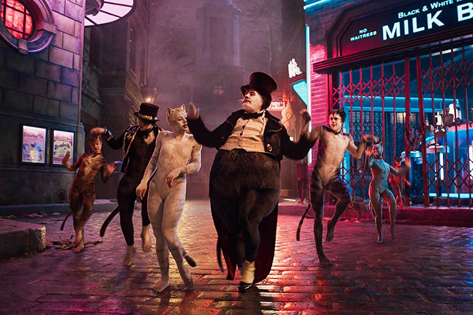 A shot of some of the cats from Cats (2019) dancing in a line. There are six of them, and they all stand and look a little like humans. They are on a cobblestone street at night. It is lit up by pink neon lights.