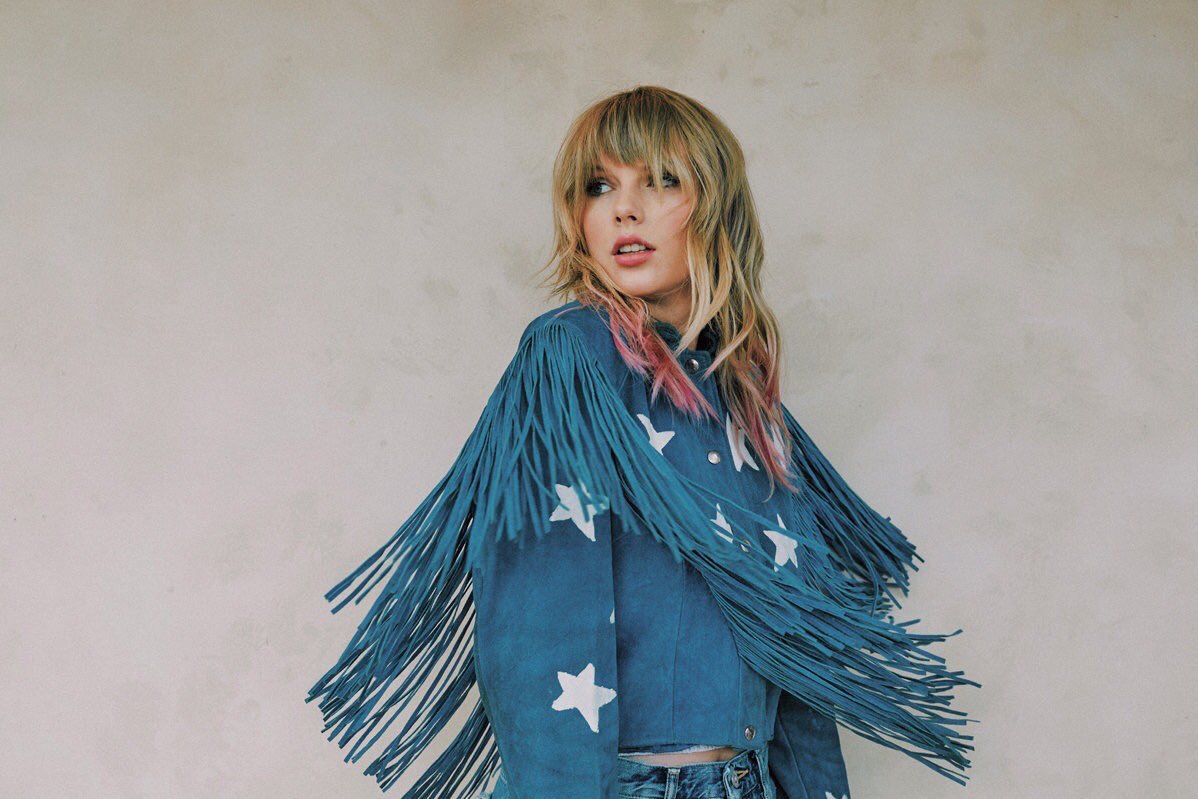 Taylor wears a blue, starry fringe jacket and twists to look over her shoulder with pink hair,
