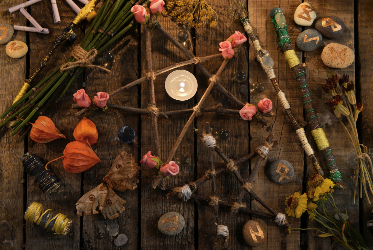 A wiccan altar contains a pentacle made out of sticks with a white candle in its center, surrounded by various flowers, stones and wands.