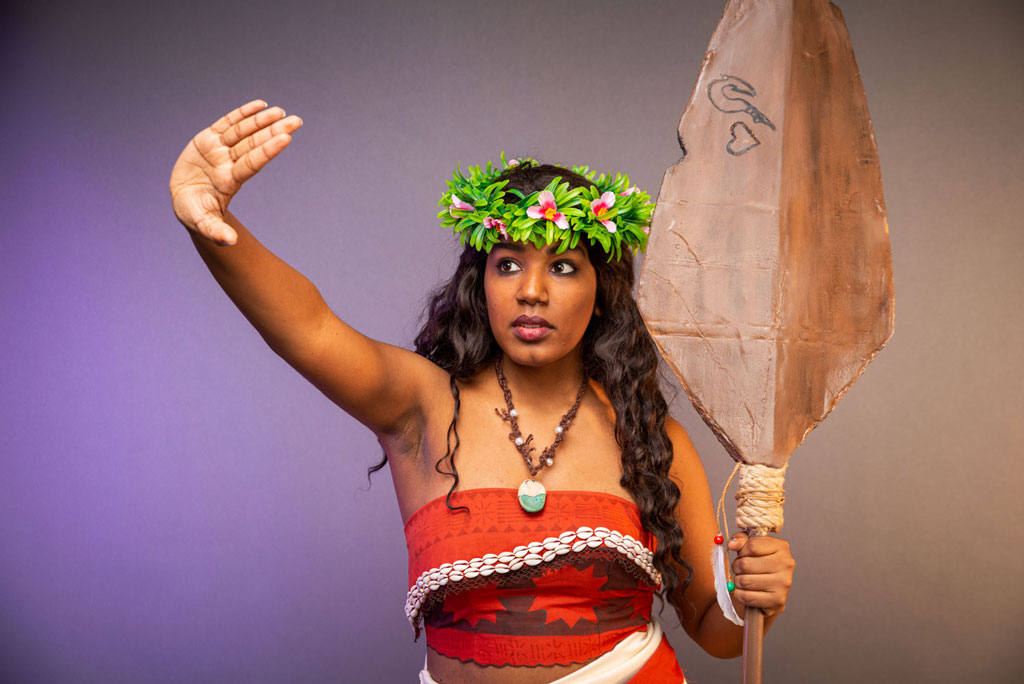 A Moana cosplayer stretches out her right arm while holding a boat oar in her left hand.