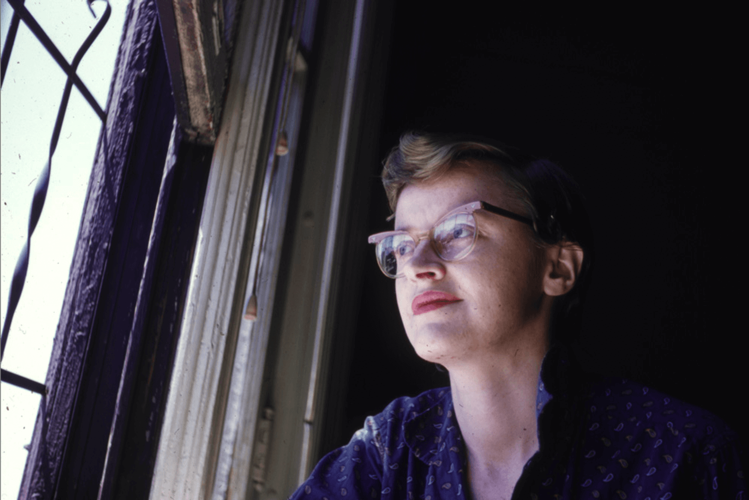 Connie Converse sits looking out a window, wearing thick, cat-eye classes and reddish lipstick. (Photo by Elizabeth Converse/The Estate of Elizabeth Converse/Squirrel Thing Recordings)