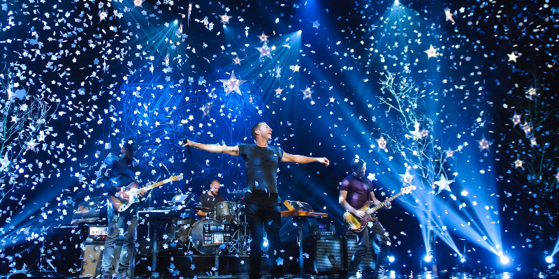 Coldplay performs their album, Ghost Stories. Chris Martin, arms spread, stands clearly in the center of the stage. The space around him is filled with confetti, hanging stars, and blue light.