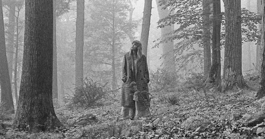 A black and white photo of Taylor Swift standing in. forest surrounded. by trees.