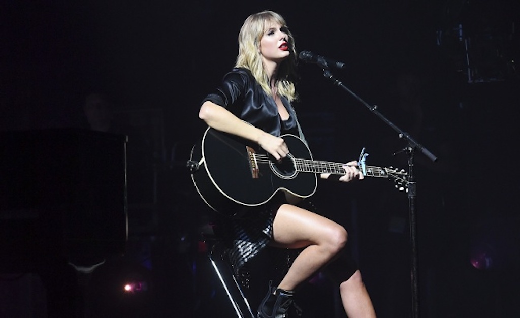 Taylor Swift sitting down at a microphone and playing the acoustic guitar.