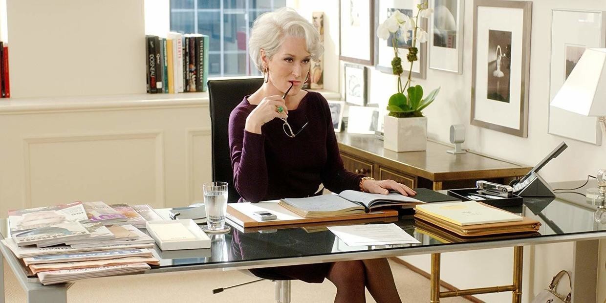 Image of Miranda Priestly seated at her desk and biting the frame of her glasses. She represents the fandom personality of the "Critic Fan."  