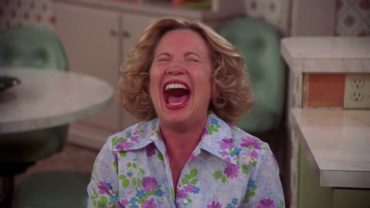 Kitty Forman is laughing and sitting at the kitchen table.