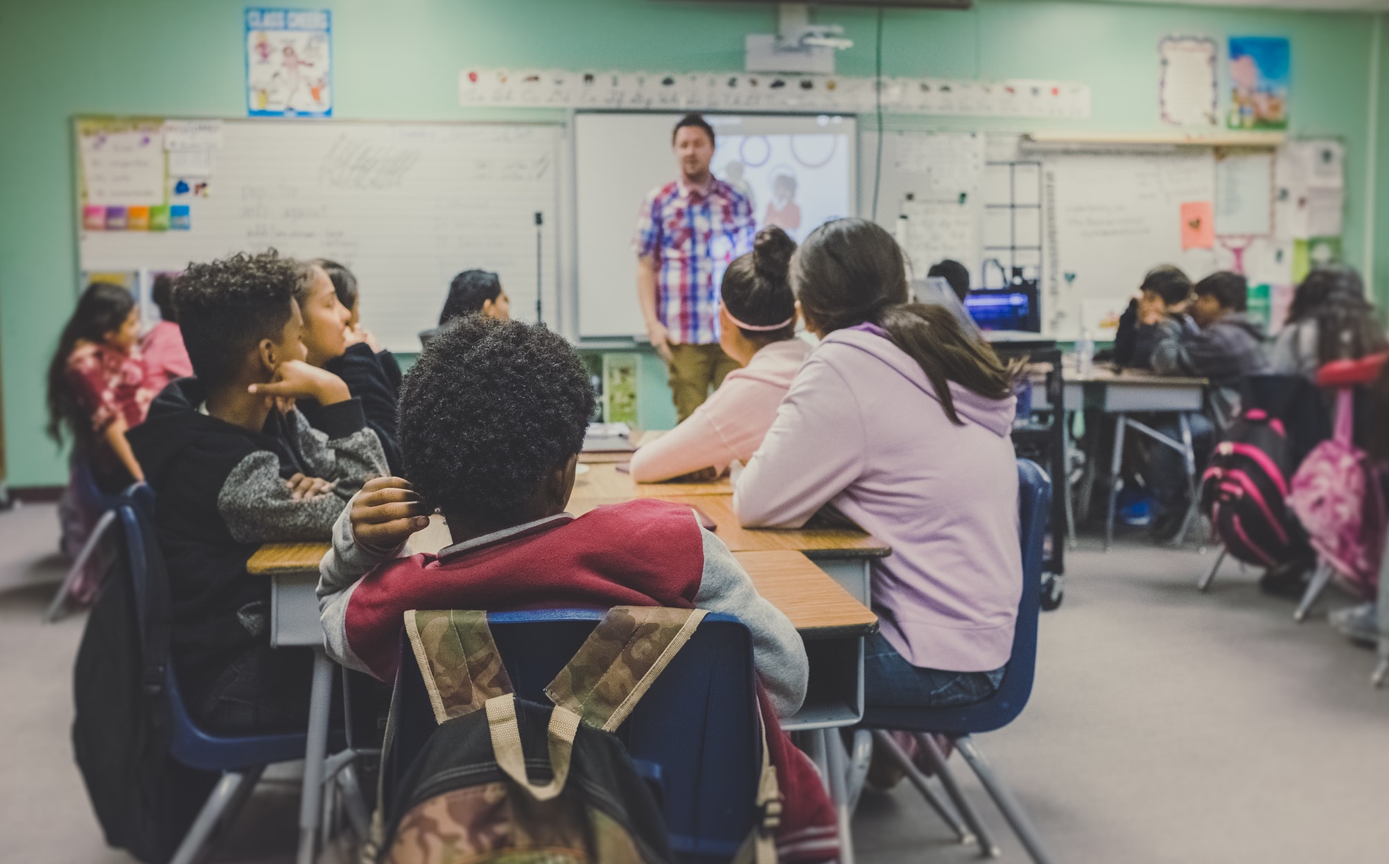 Students sitting at desks in a classroom, watching their teacher.