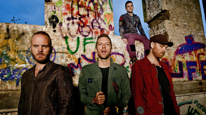 Will Champion, Chris Martin, and Jonny Buckland stand in front of a graffitied wall, decorated for their "Mylo Xyloto" album. Guy Berryman sits on the wall.