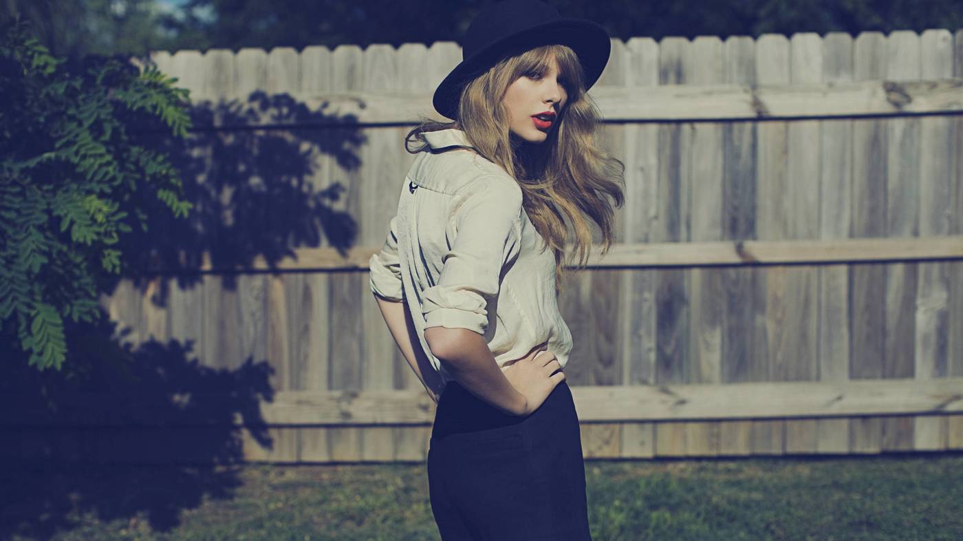 Taylor Swift standing with her hands on her hips in front of a wooden fence.