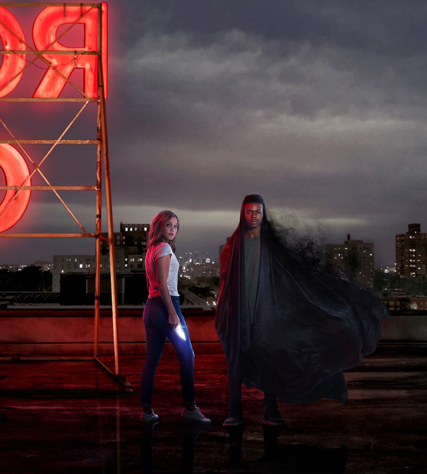 Tandy, a girl with a dagger of glowing light, and Tyrone, a boy with a jacket that fades into shadowy mist, stand on a roof-top near a water tank against a dark, cloudy sky. (Photo by Freeform.)