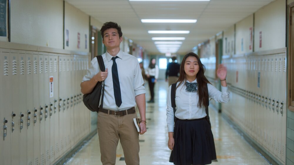 Jamie and Eli standing in the middle of a school hallway. There are lockers lining the walls Jamie is waving 