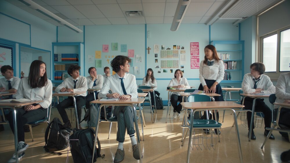 Jamie standing by her desk in a classroom. The rest of class is seated and  looking at her as she introduces herself