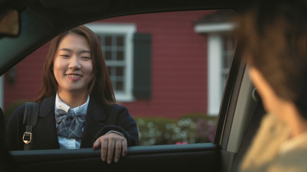 Scene from Parked in America; Jamie standing at car window, smiling at the driver. The driver's back of head is on the right side of screen, blurry. 