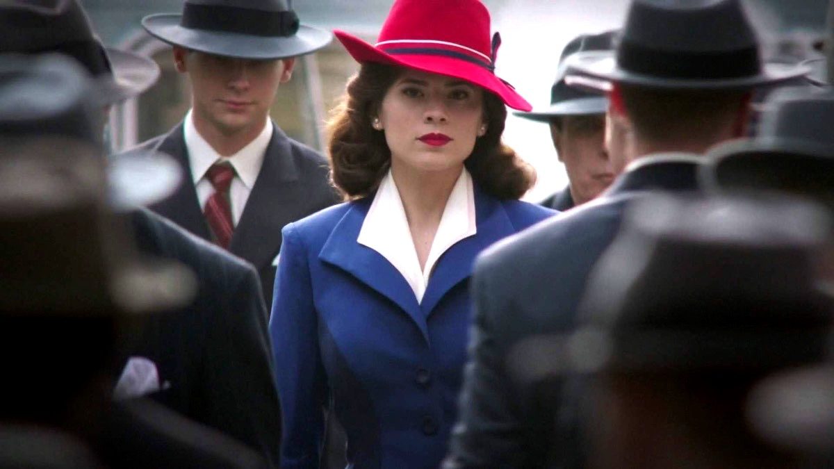 A promotional image for one of the first Marvel shows to spin off of the MCU, Agent Carter, showing Peggy in a navy pantsuit and bright red hat walking through a sea of men in grey and black suits. (Photo by ABC and Marvel.)