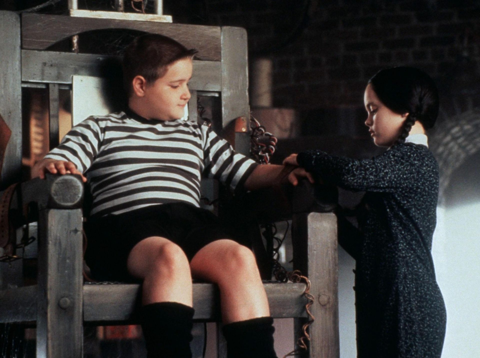 Pugsley sits in a makeshift electric chair while Wednesday straps him in. 