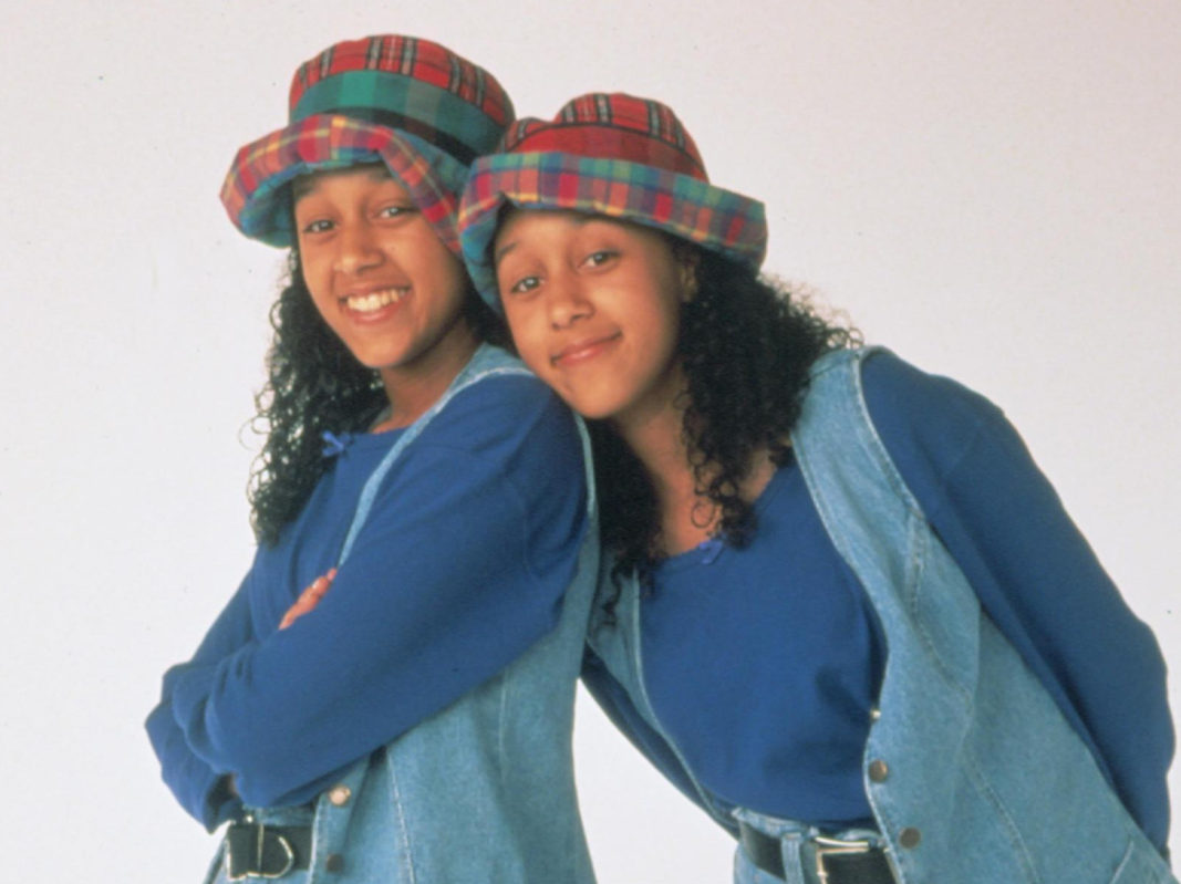 Against a white backdrop, Tia and Tamara pose leaning on each other while wearing matching outfits: plaid hats, jeans, denim bests, and blue shirts. 