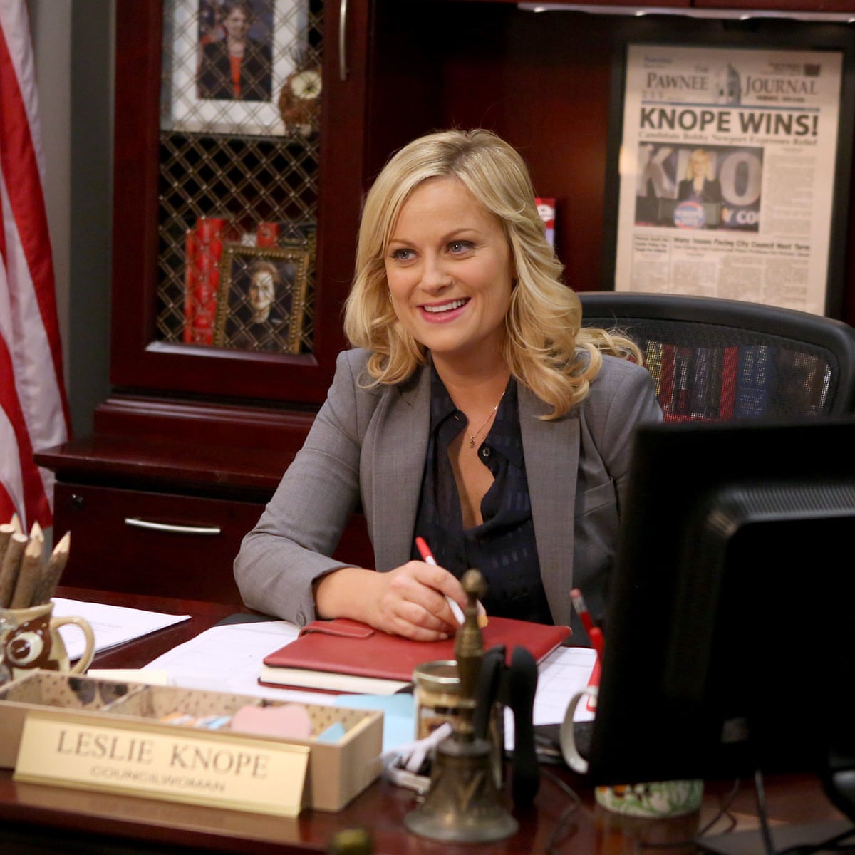 Leslie Knope sits at her large office desk, pen in hand and smiling. 