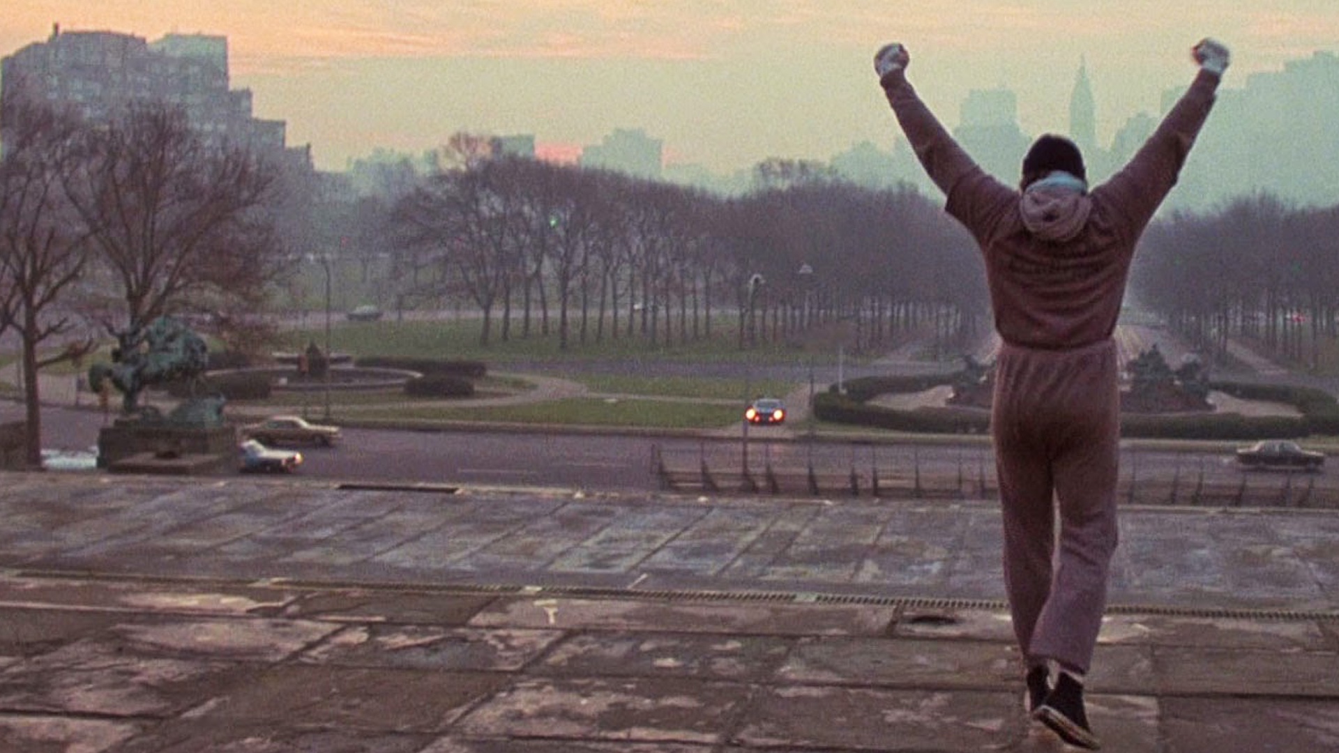 Rocky Balboa, in grey training sweats, stands with both arms held above his head in victory as he looks down the stairs at the city of Philadelphia.