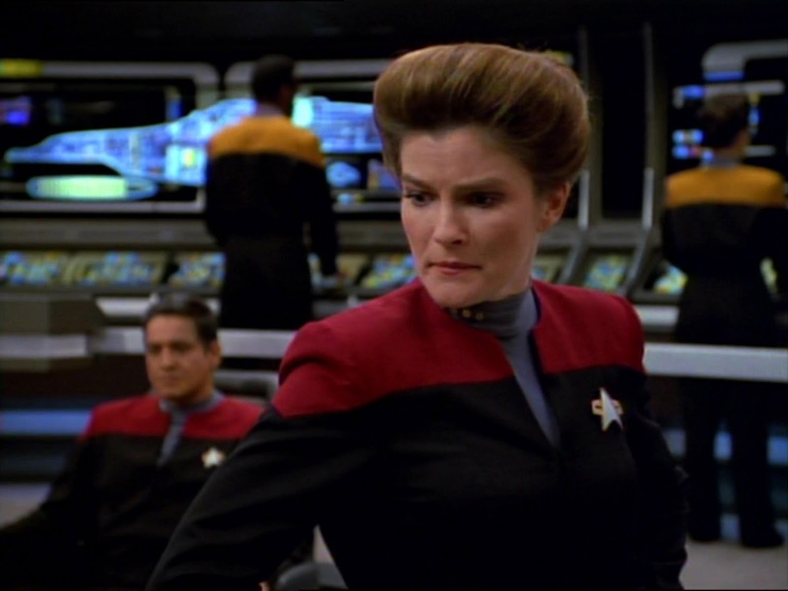 Captain Janeway stares intently while standing on the starship's bridge. She is wearing her black and red uniform complete with the Starfleet insignia communication badge. 
